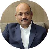 Man with a mustache wearing a dark suit jacket and white shirt sitting on a beige couch.